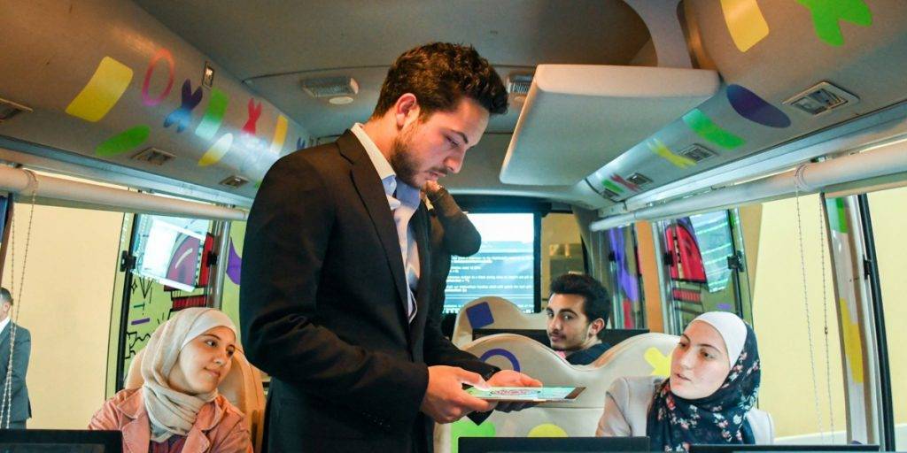 Crown Prince of Jordan in a suit hands a tablet to a woman in a hijab on a bus, while two other young adults, also in hijabs, watch. The bus, featuring colorful decorations and computer monitors, highlights innovations shaping the MENA region.