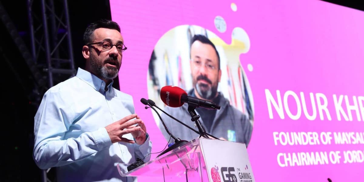 A man speaks at a clear podium with microphones, while a large screen behind him displays his image and the text Nour Kheder, Founder of Maxsmith and Maysalward, notable for his success in the gaming industry, Chairman of Jordan.